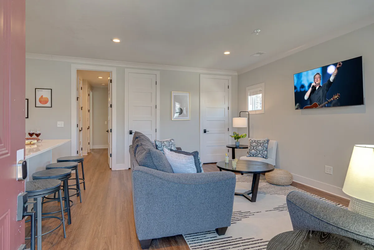 A contemporary open-concept living room featuring a comfortable sofa, coffee table, wall-mounted television, bar stools at the kitchen counter, and stylish artwork. The layout encourages relaxation and socializing, with easy sightlines into the adjacent kitchen area. Warm lighting and modern decor create a cozy yet vibrant atmosphere ideal for group gatherings.