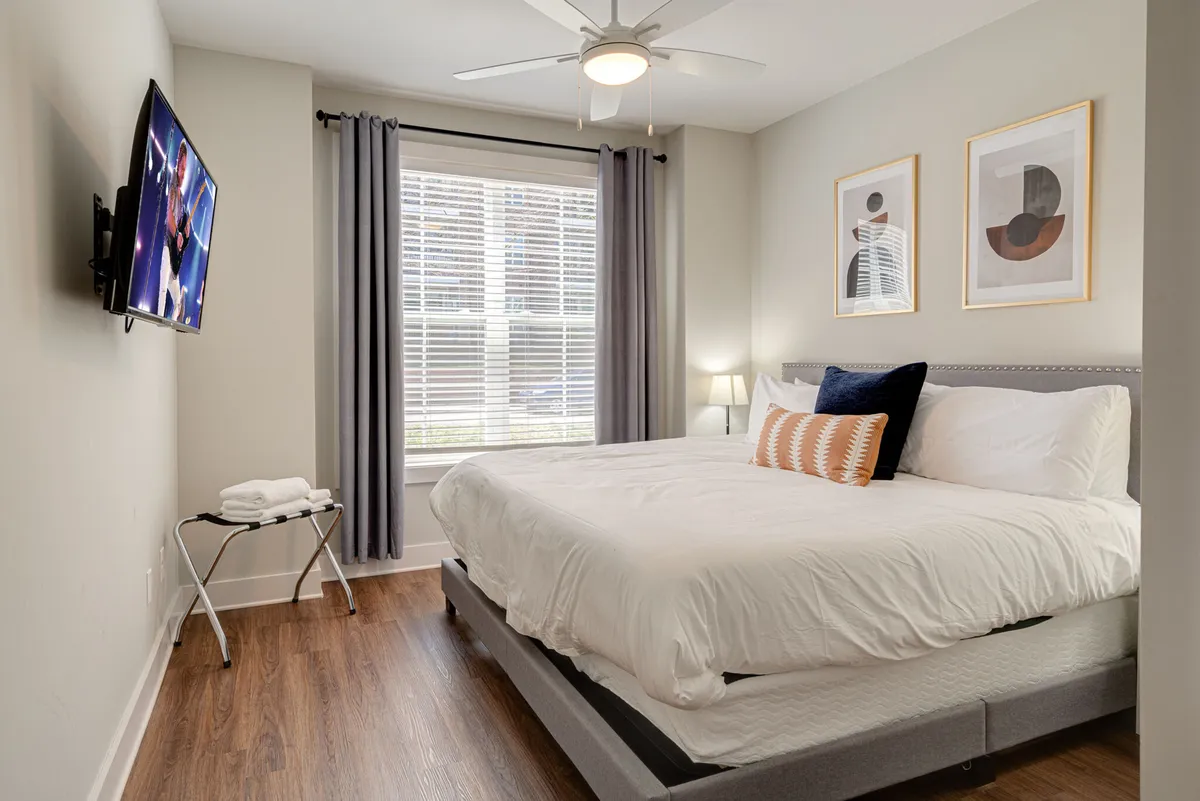 A tranquil bedroom with a king-sized bed, a Smart TV mounted opposite, a window allowing natural light, and calming artwork on the wall. The scene is designed for relaxation, whether watching TV, reading, or napping.