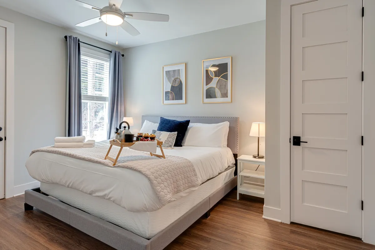 A bright bedroom featuring a plush king-sized bed, soft window curtains letting in natural daylight, a tray with breakfast items on the bed, and thoughtfully chosen wall art. The space feels private and inviting, ideal for restful nights or lazy mornings.