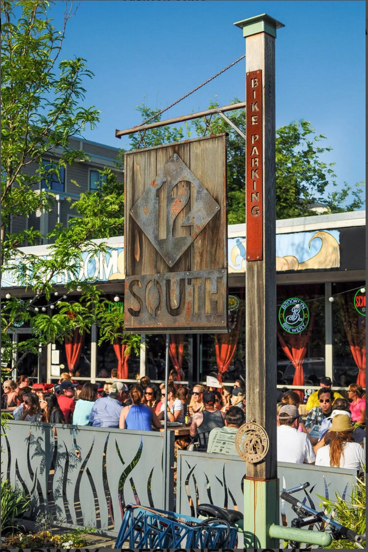 A bustling streetscape in Nashville's Melrose/12 South area, this image features the '12 South' sign, a diverse crowd of people dining outdoors, and a bike parking sign, showcasing the neighborhood's lively social energy and pedestrian-friendly vibe.